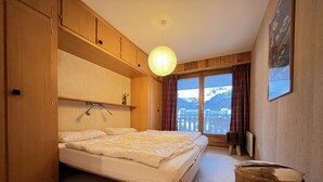 Ein Schlafzimmer mit einem Bett, einem Holzschrank, einer Pendelleuchte, einem kleinen Hocker und einem Fenster mit Bergblick.