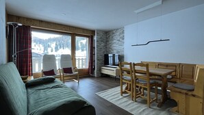 Ein modernes Wohnzimmer mit einer Couch, einem Holzesstisch, Stühlen, einem Fernseher und einem großen Fenster mit Blick auf eine verschneite Landschaft.