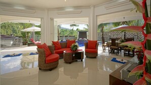 A spacious indoor area with a pool, red and brown wicker sofas, a dining table, and tropical plants. - Luxurious, quiet, private Pool-Villa Lotus, 7/7 housekeeper/butler (Phuket)