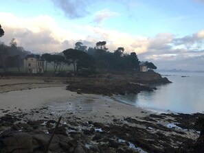A coastal landscape with a sandy beach, rocky shoreline, and a building with a fence in the foreground.