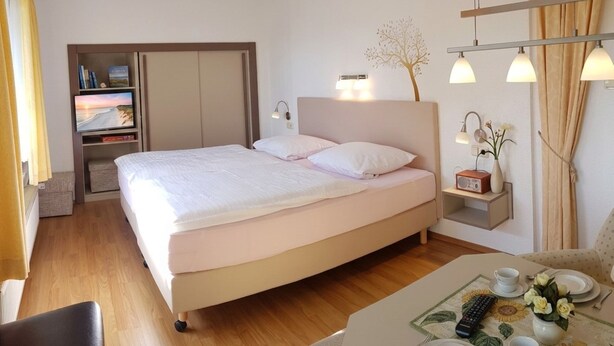 Habitación de hotel con cama, mesita de noche, lámpara y una pequeña mesa con un jarrón y flores.