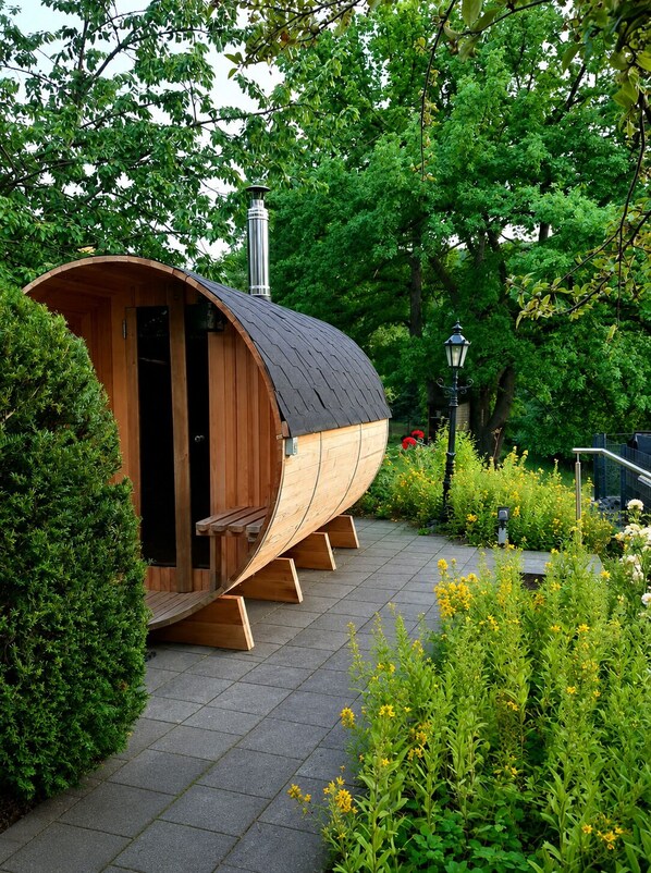A wooden sauna structure with a curved roof, surrounded by greenery and a paved walkway.