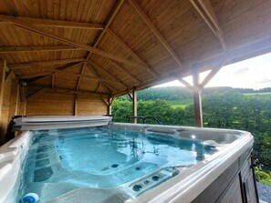A hot tub under a wooden roof with a view of a green landscape.