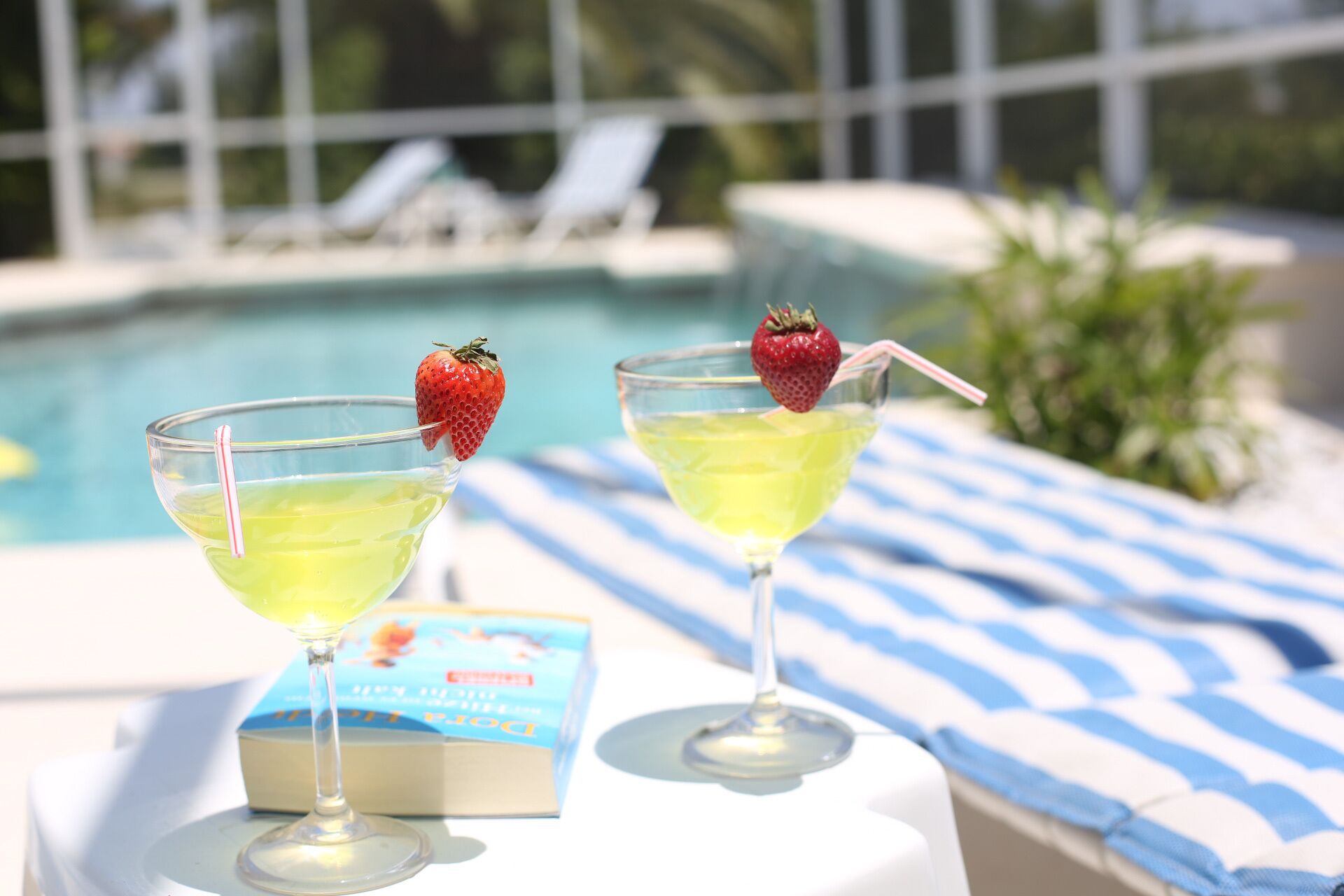 Two martini glasses with yellow drinks and strawberries on a table near a pool with lounge chairs and a book titled "Dora".