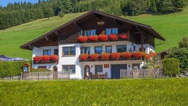 A two-story chalet with a balcony, surrounded by a fenced property and a forested hillside. - Apartment A (2-3) - Apartments and Bed & Breakfast Eberle (Jungholz)