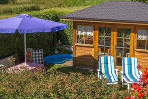 A wooden gazebo with a pool, surrounded by lush greenery and a patio with a table and chairs.