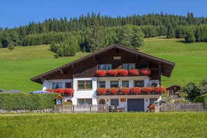A two-story building with a balcony adorned with red flowers, surrounded by a green landscape and a forest in the background. - Apartment A (2-3) - Apartments and Bed & Breakfast Eberle (Jungholz)
