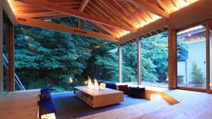 A modern interior with a wooden ceiling, a central fireplace, and large windows offering a view of a forested area.