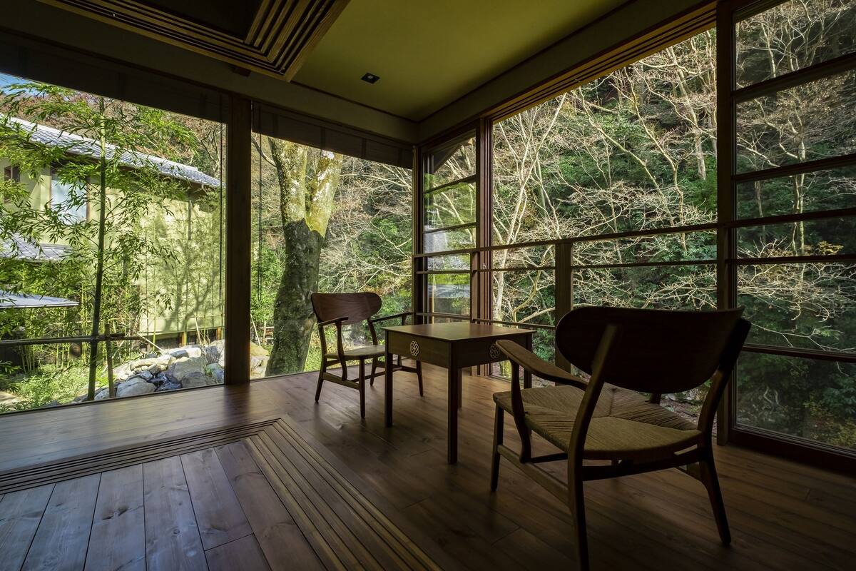 Amplia habitación con ventanas grandes que ofrecen vista a un área boscosa. Hay una mesa de madera y dos sillas.