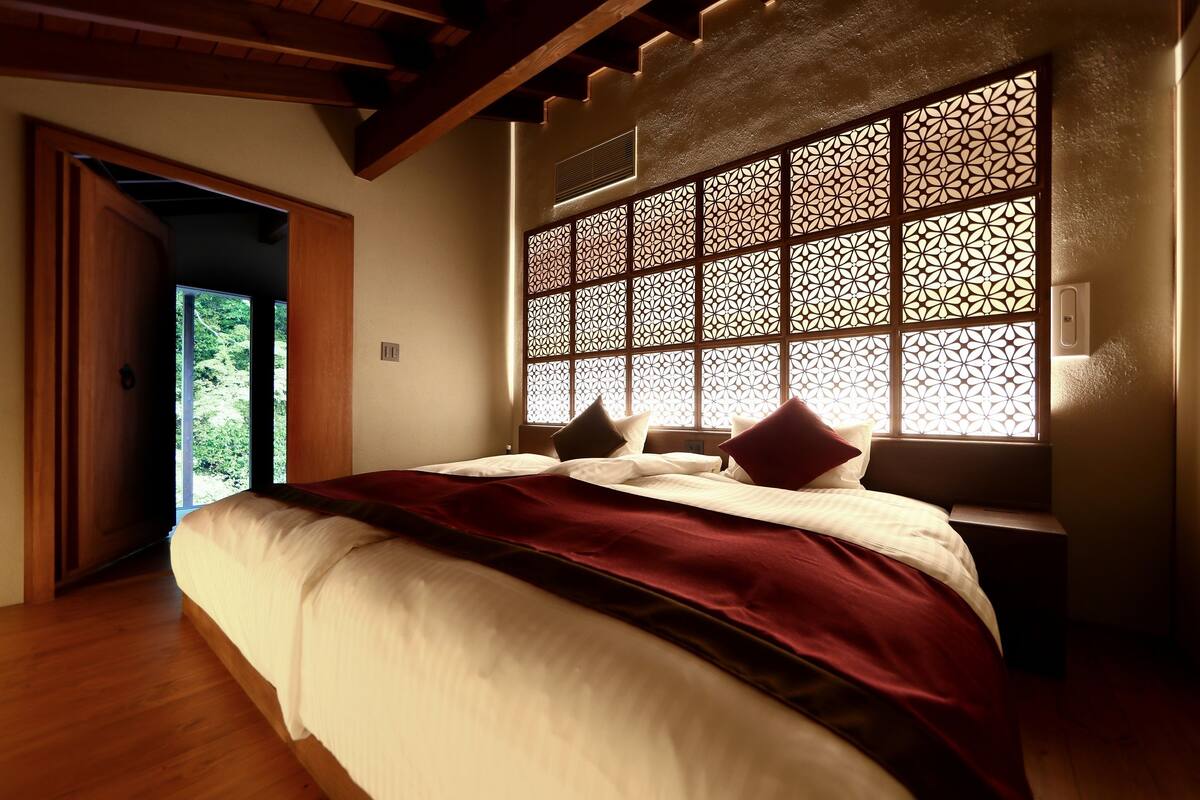 Un dormitorio con una cama grande, suelo de madera, ventana decorativa con un diseño geométrico y vistas al exterior con vegetación.