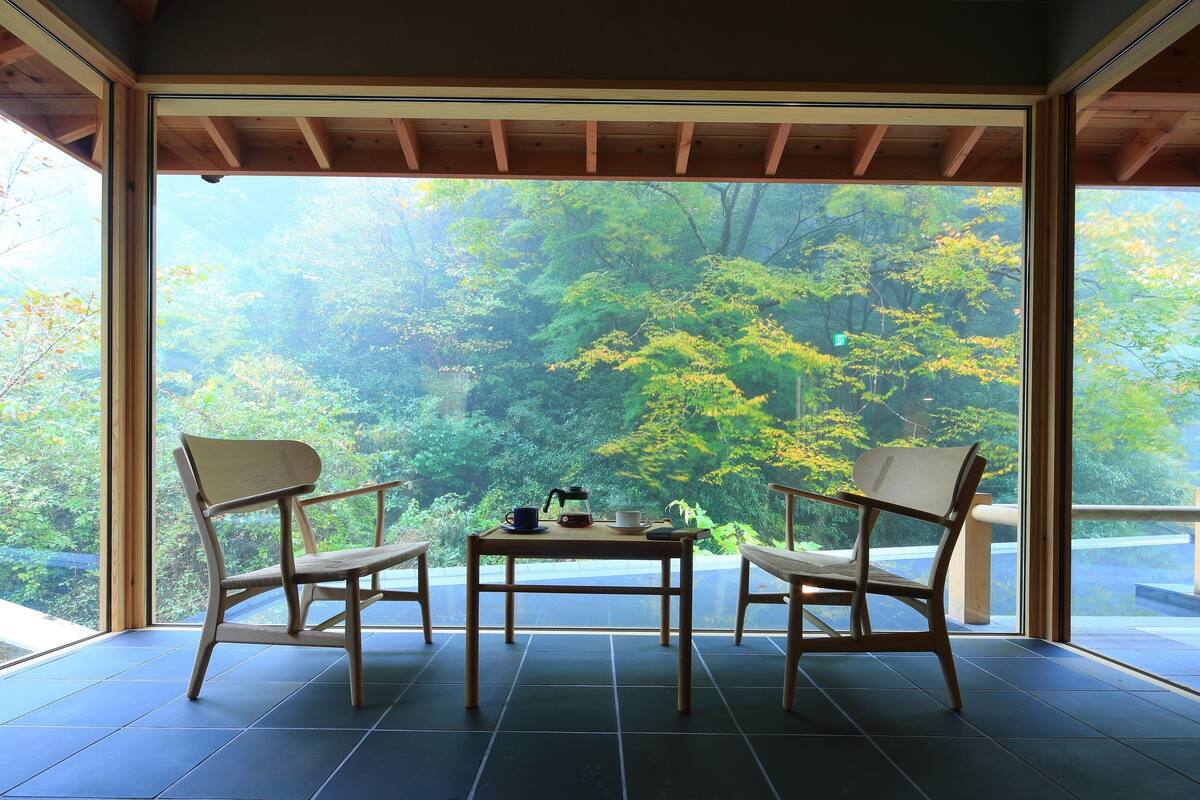 Una habitación con un ventanal grande con vistas a un bosque, dos sillas, una mesita con un hervidor y tazas, y un piso de baldosas.