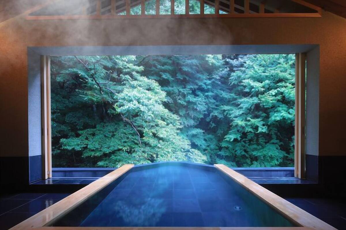 Un área de spa con un amplio ventanal con vistas a un bosque, una bañera de hidromasaje y una estructura de techo de madera.