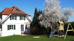 Ein zweistöckiges Haus mit rotem Ziegeldach, einem hölzernen Spielhäuschen mit Rutsche und einem großen, blühenden Baum.