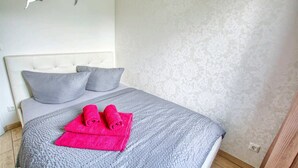 Un llit ben fet amb roba de llit gris i dos coixins, un parell de tovalloles rosa brillant a sobre del llit, i una paret amb un disseny floral discret.
