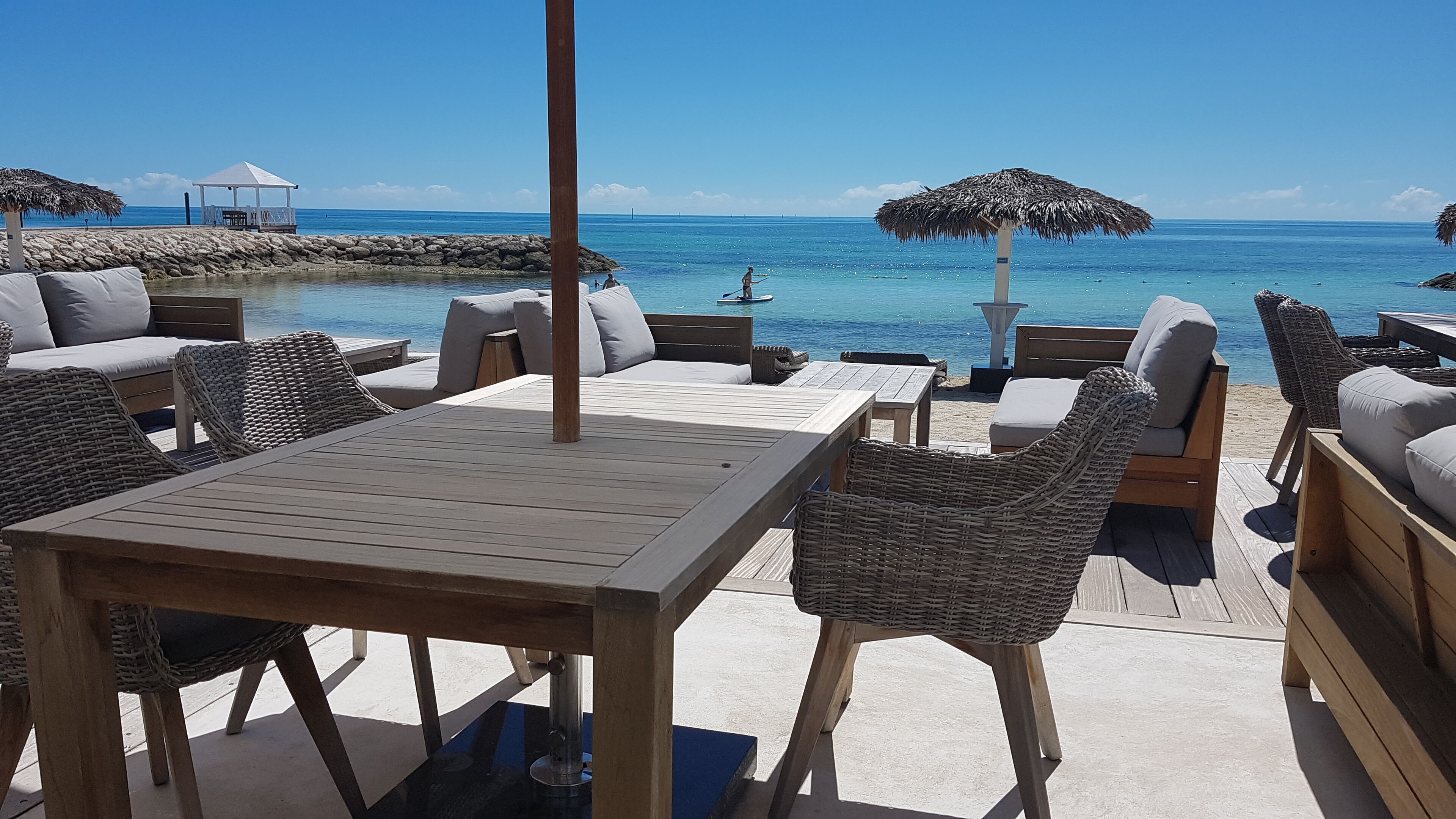 A wooden deck with wicker furniture and a table, overlooking a clear blue sea.