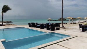 A pool area with lounge chairs and umbrellas, a clear view of the ocean, and a sandy beach in the background.