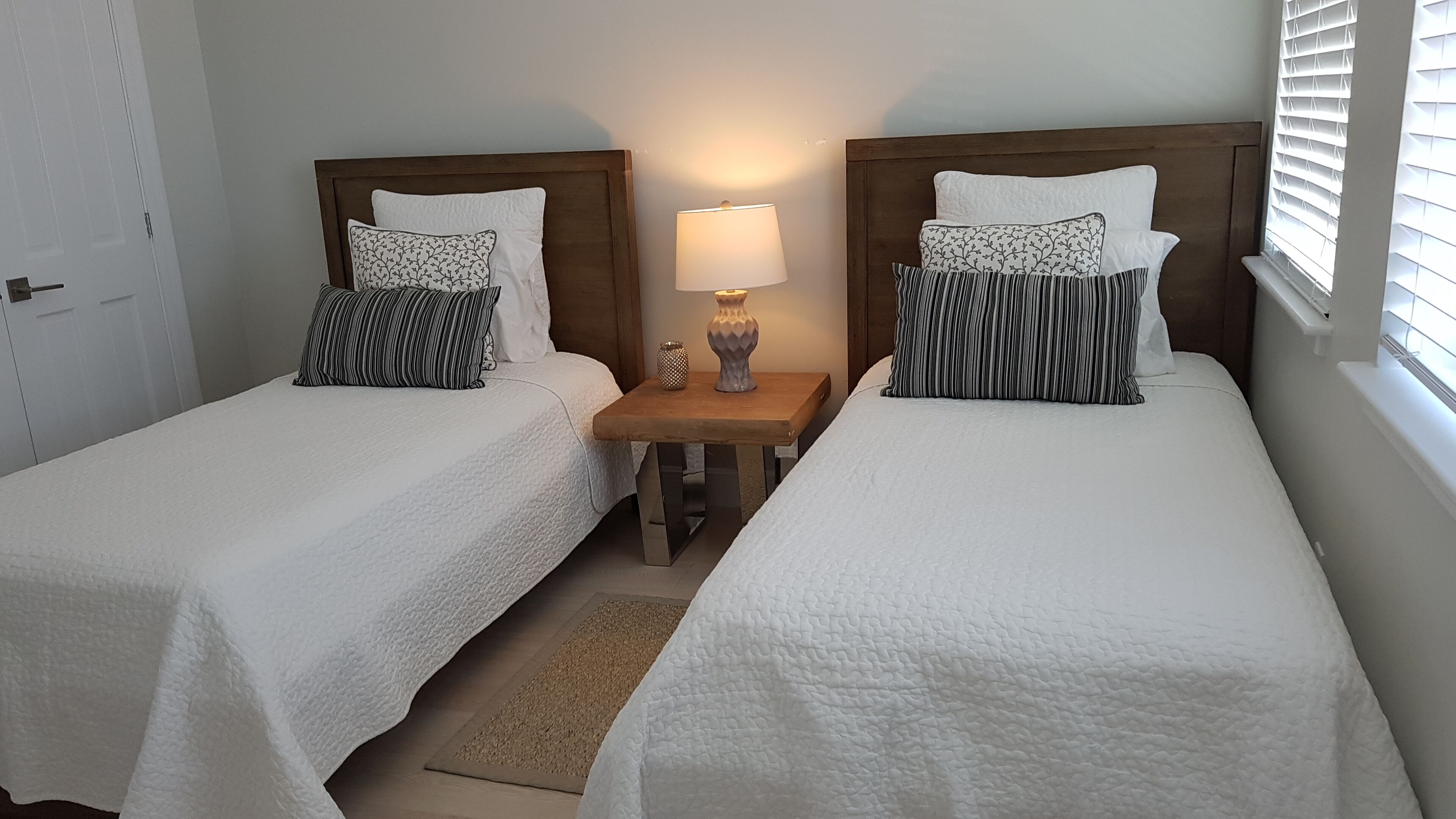 A hotel room with two single beds, a nightstand with a lamp, and a window with blinds.