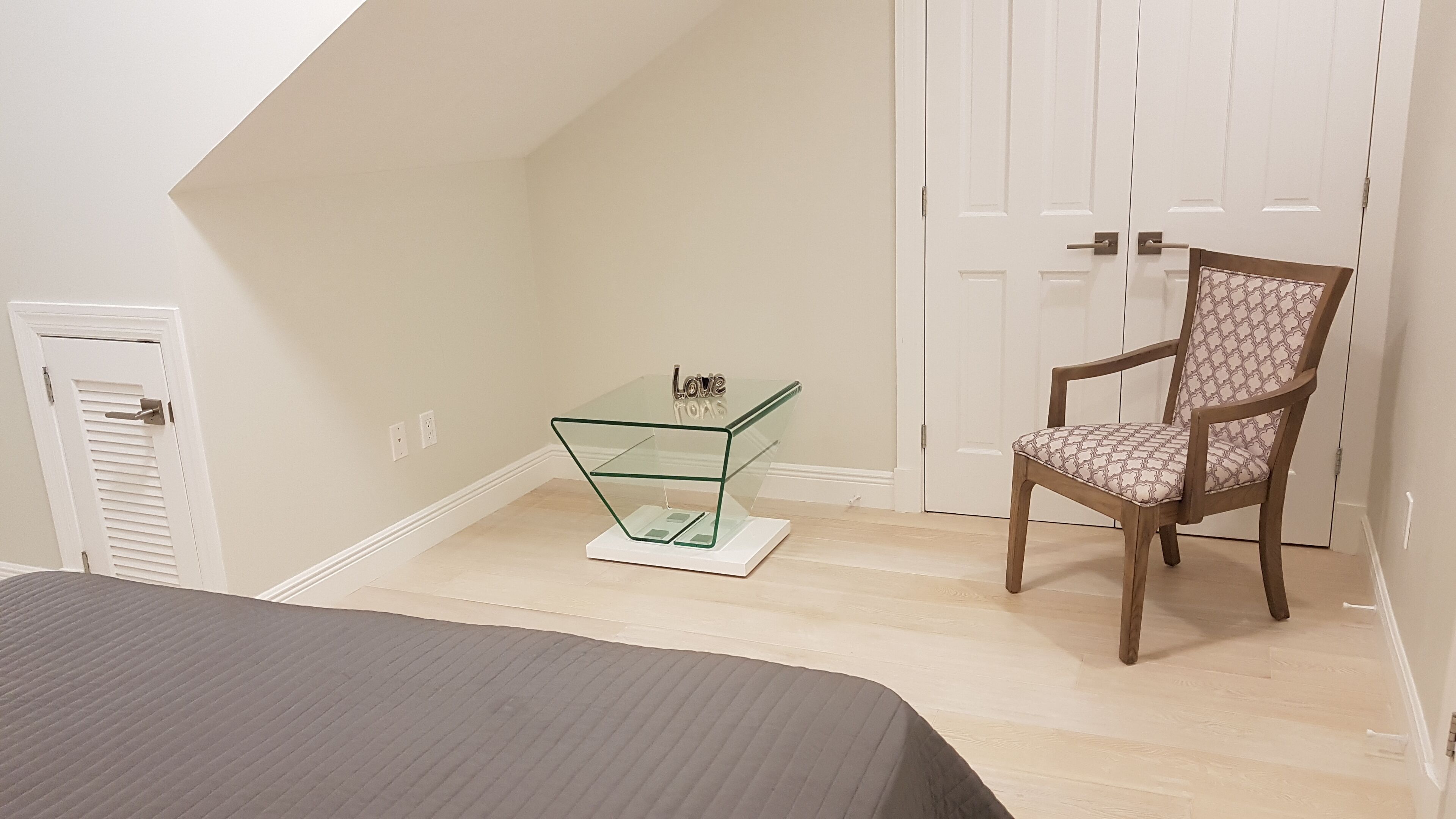 Un dormitorio con una cama, una mesita de cristal con un objeto decorativo, una silla de madera y una puerta blanca.