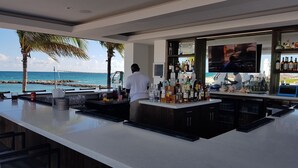 A bar with a view of the ocean, a bartender, and various bottles and glasses.