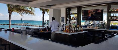 A bar with a view of the ocean, a bartender, and various bottles and glasses.