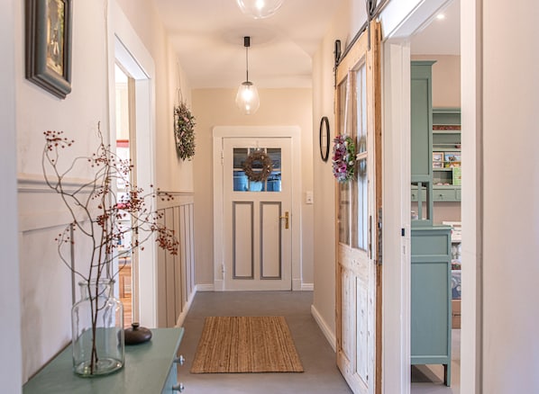 A hallway with a door, a hanging light fixture, a rug, and a vase with branches.