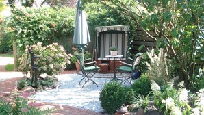 Un patio con una silla exterior a rayas, una mesa con una planta en maceta y un jardín con diversas plantas y una estatua decorativa de un pájaro.