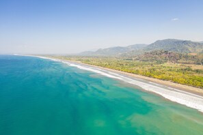 Eine Luftaufnahme einer Küstenlandschaft mit Strand, klarem türkisblauem Wasser und einer bewaldeten Hügelskette.