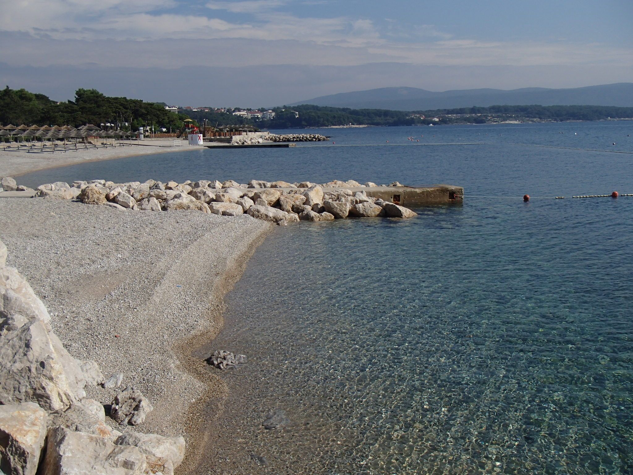 A pebble beach with clear water, a rocky breakwater, and a distant shoreline with buildings.