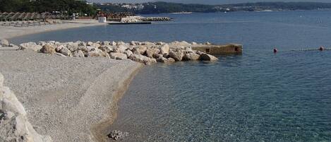 A pebble beach with clear water, a rocky breakwater, and a distant shoreline with buildings.