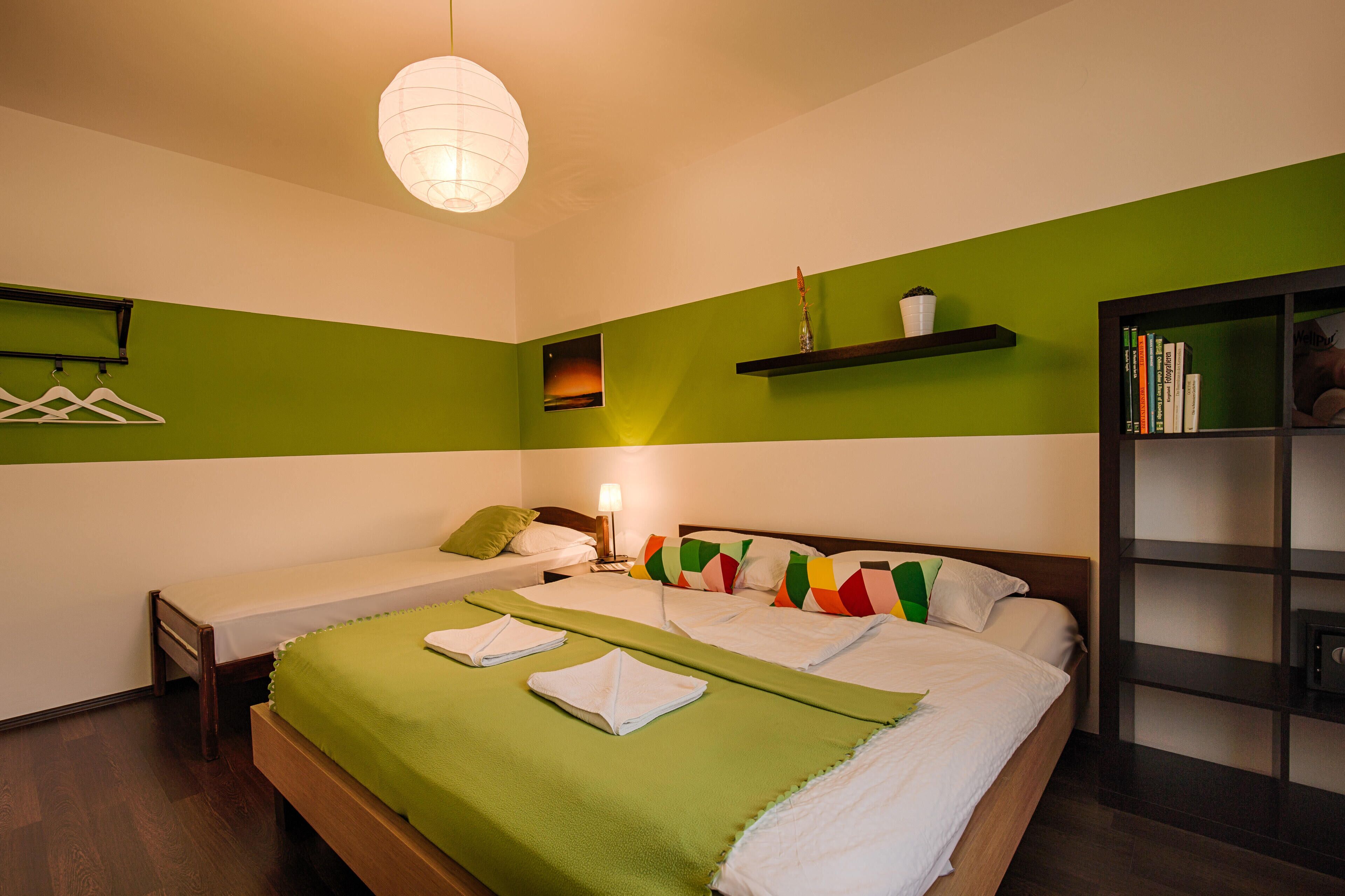 A hotel room with a bed, a bedside table, a lamp, a shelf with books, and a wall-mounted coat rack.