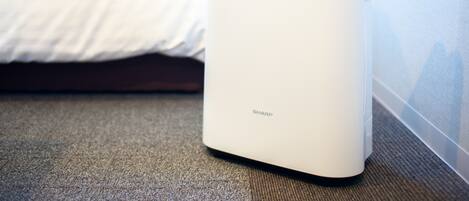 A white air purifier with the brand name 'SHARP' on a carpeted floor next to a bed with white bedding.