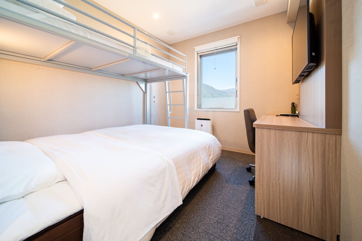 A compact hotel room with a bunk bed, a desk, a chair, a TV, and a window offering a view of the outdoors.