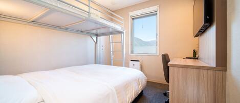 A compact hotel room with a bunk bed, a desk, a chair, a TV, and a window offering a view of the outdoors.