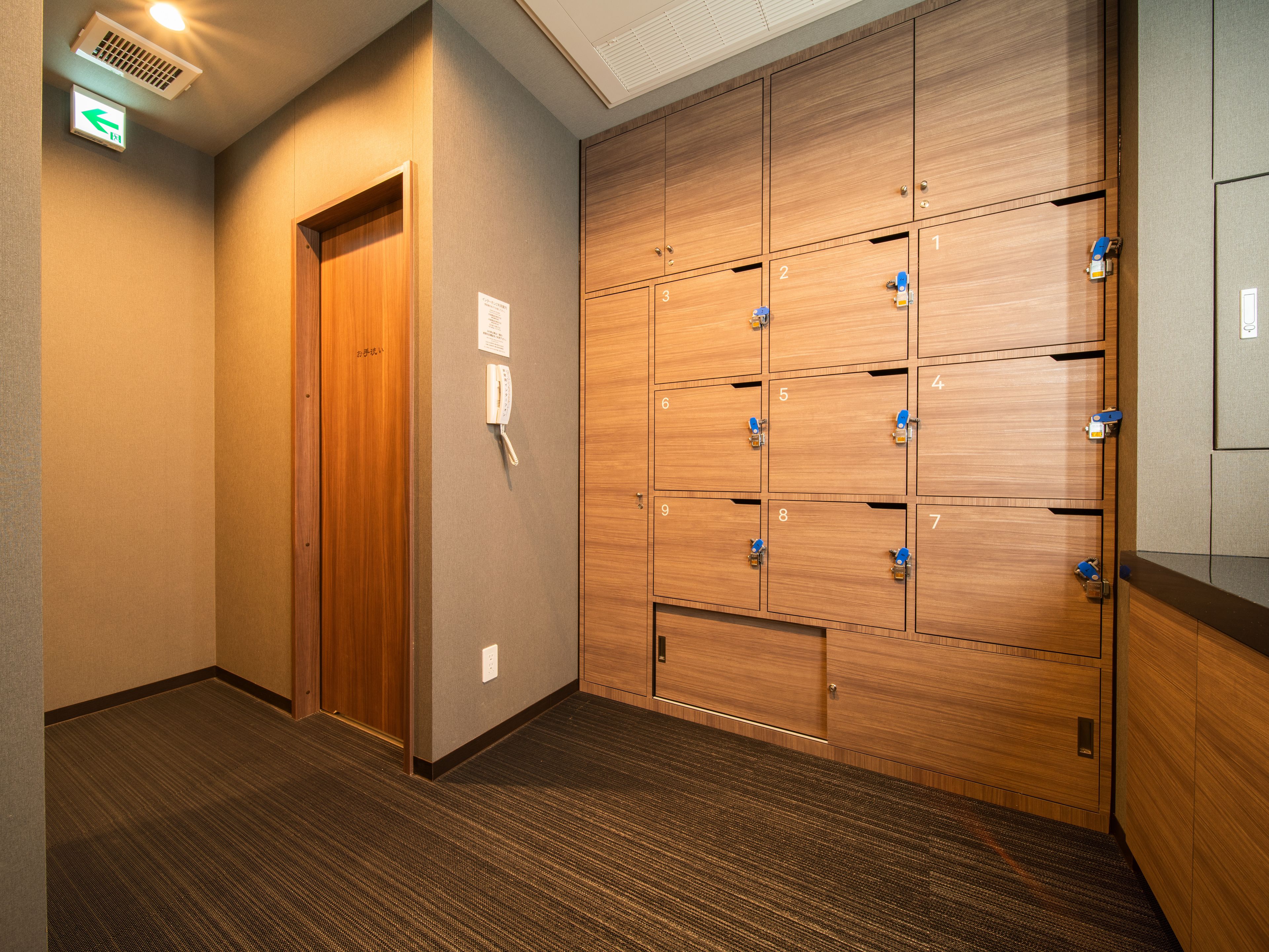 A hotel hallway with a wooden door, a wall-mounted phone, and a row of numbered lockers.