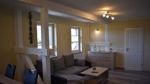 Ein Wohnzimmer mit Sofa, Couchtisch und einem Essbereich mit Stühlen. Es gibt eine Küchenzeile mit Schrank und einer Tür.