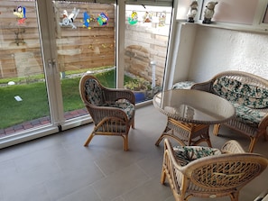 Eine Terrassenfläche mit Korbmöbeln, eine Glastür, die in einen Garten führt, und eine kleine Ablage mit Dekoration.