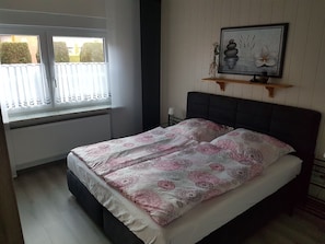 Ein Schlafzimmer mit einem Bett, einem Fenster mit Vorhängen, einem Heizkörper, einer Wandablage mit Dekoration und einem gerahmten Bild an der Wand.