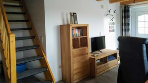 Een houten trap die leidt naar een woonruimte met een televisie, een boekenkast en een stoel.