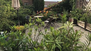 Eine Holzterrasse mit Gartenmöbeln, umgeben von üppigem Grün und einem Backsteinhaus im Hintergrund.