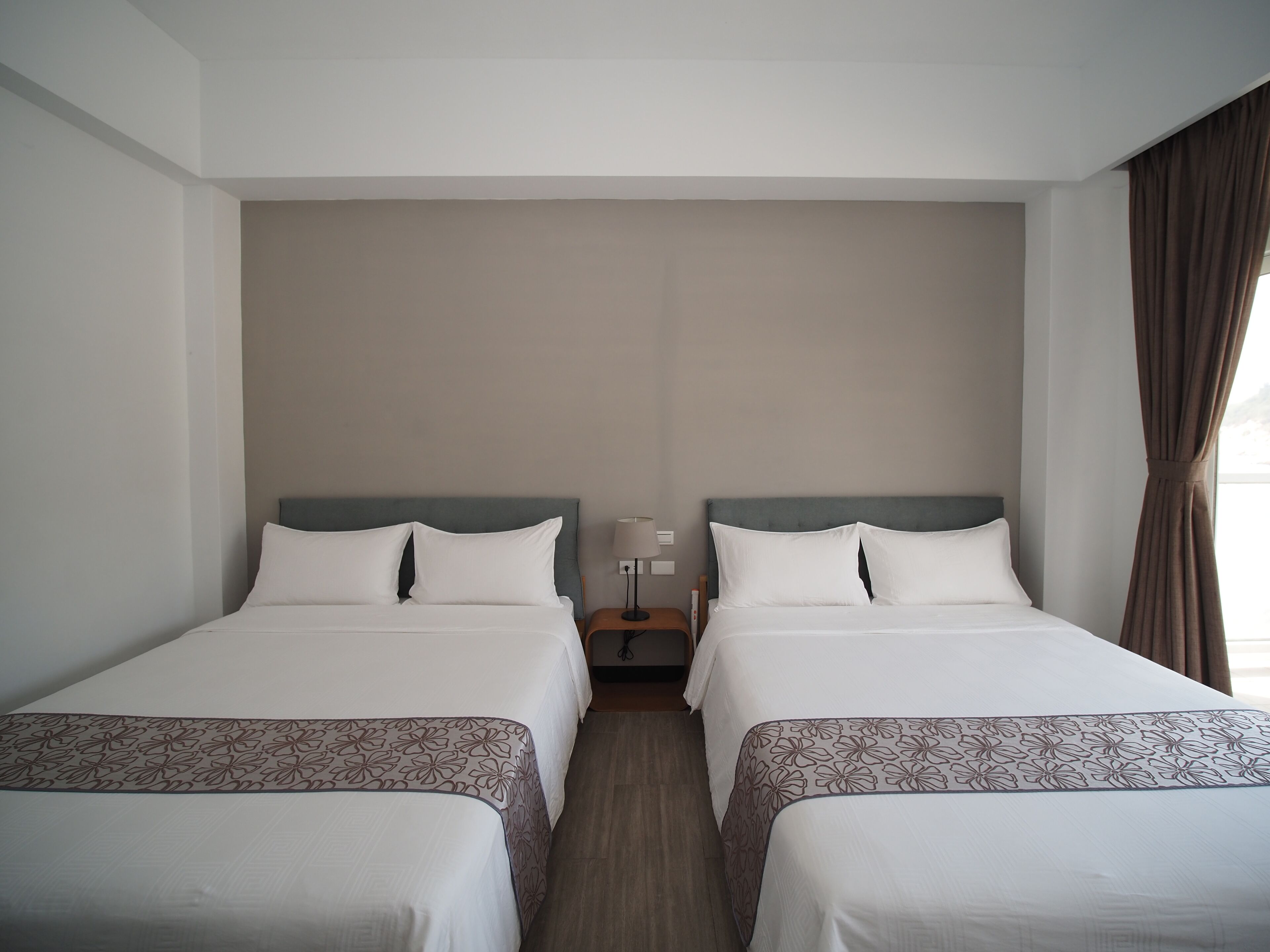 A hotel room with two single beds, a nightstand with a lamp, and a window with curtains.