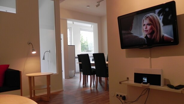 Ett modernt vardagsrum med en platt-TV som är monterad på väggen, en matplats med bord och stolar samt ett kök i bakgrunden.