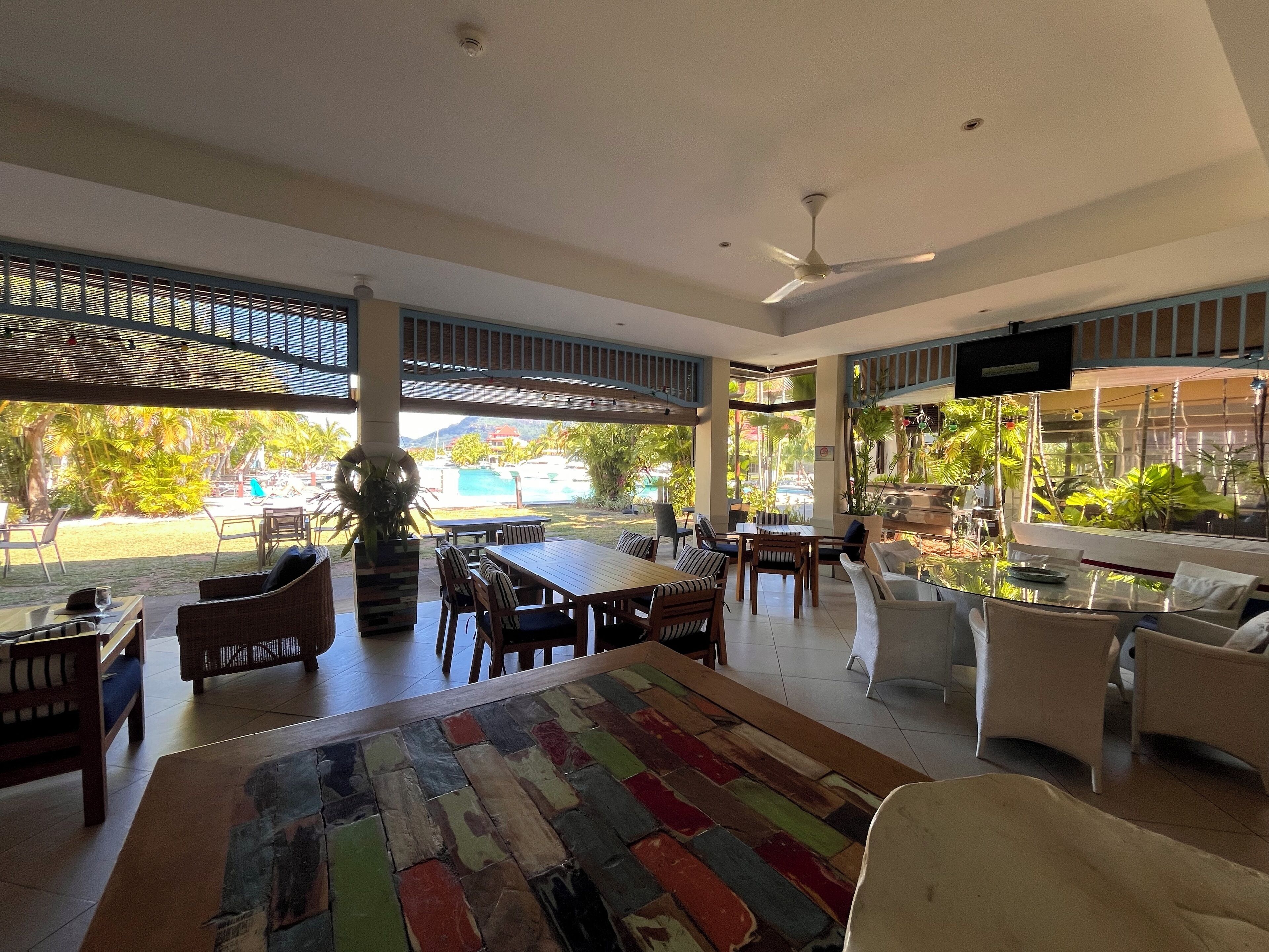 A spacious indoor area with a large wooden table, several chairs, and a colorful rug. There is a ceiling fan and a view of a pool and greenery outside.