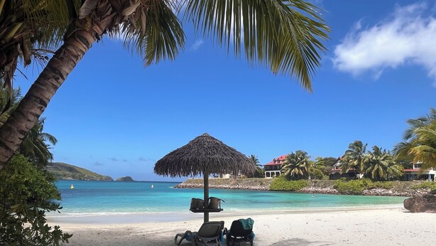 Une plage avec un parasol en chaume, des chaises longues et une vue dégagée sur l’océan et les bâtiments situés au loin.