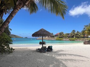 Ein Strand mit einem Reetdachschirm, Liegestühlen und freiem Blick auf das Meer und entfernte Gebäude.