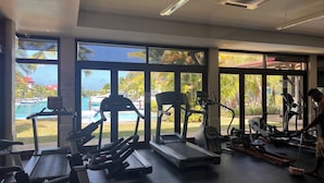 Une salle de sport équipée de divers appareils d’exercice, avec de grandes fenêtres donnant sur une piscine et des palmiers.