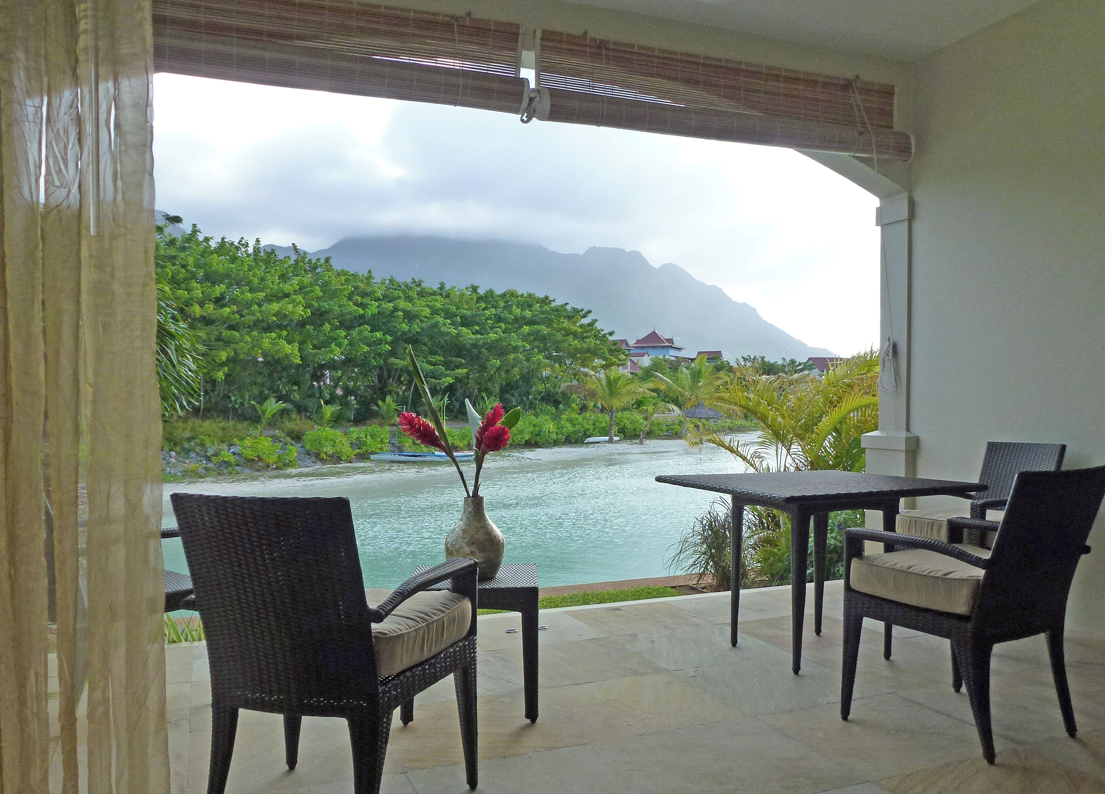 A room with a view of a body of water, greenery, and mountains. There is a table and chairs set up for dining.