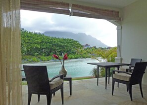 A room with a view of a body of water, greenery, and mountains. There is a table and chairs set up for dining.