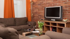 A living room with a brick wall, a brown sofa, a glass coffee table, a flat-screen TV, a floor lamp, and a bookshelf.