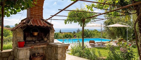 Cocina exterior de piedra con chimenea, piscina y vistas al mar.