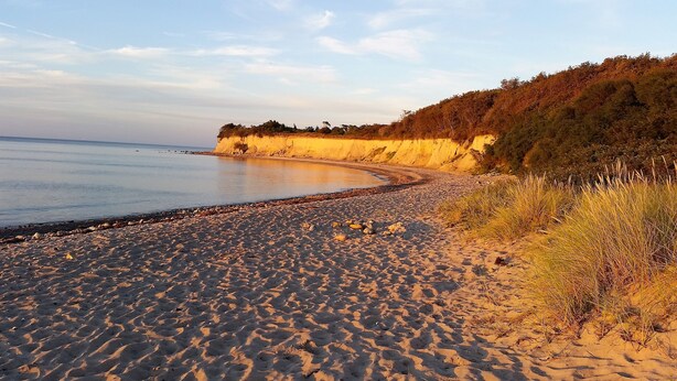 A sandy beach with a curved shoreline, a grassy cliff, and a calm sea.